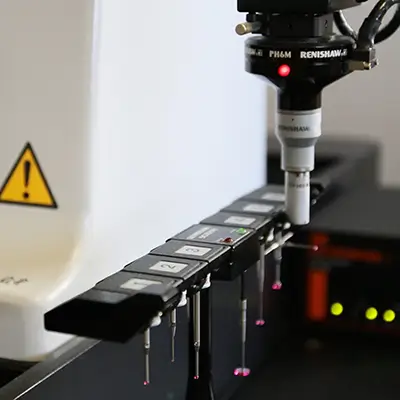 Nahaufnahme einer Präzisionsmessmaschine mit einem Renishaw-Tastkopf, die eine Reihe von beschrifteten Metallteilen prüft oder kalibriert, wobei im Hintergrund ein Warnschild zu sehen ist.